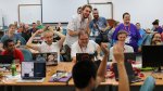 Une trentaine de personnes assises devant des ordinateurs portables rient. Une personne au premier plan de dos applaudit.