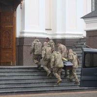 Six hommes en uniforme, vus de dos, portent à cercueil, d'une voiture à un bâtiment dont les portes sont ouvertes.